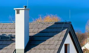 Dachówka i komin z elementami instalacji odgromowej. Niebo błękitne, drzewa w tle.