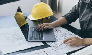 Architekt pracuje nad projektem, analizując plany i obliczając powierzchnię całkowitą budynku. Na biurku leży żółty kask.