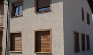 Szara, chropowata struktura elewacyjna domu z brązowymi ramami okiennymi i roletami.
