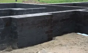 Fundamenty domu, ściany z cegły pokryte czarną izolacją. Widać prace budowlane, ziemia i gruz.