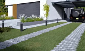 Nowoczesny podjazd z kostki bauma, z zieloną trawą i białymi kwiatami, prowadzi do zadaszonego miejsca parkingowego, gdzie stoi czarny SUV.