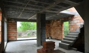 Wnętrze budowy z widocznym stropem gęstożebrowym, schodami, ceglanymi ścianami i otwartymi drzwiami na zewnątrz.