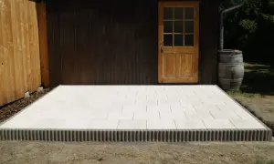 Nowy taras betonowy z jasnych płyt, otoczony ozdobnym krawężnikiem, przy drewnianej szopie z beczką na deszczówkę.
