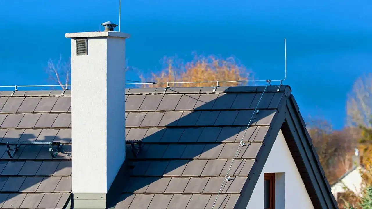Dachówka i komin z elementami instalacji odgromowej. Niebo błękitne, drzewa w tle.
