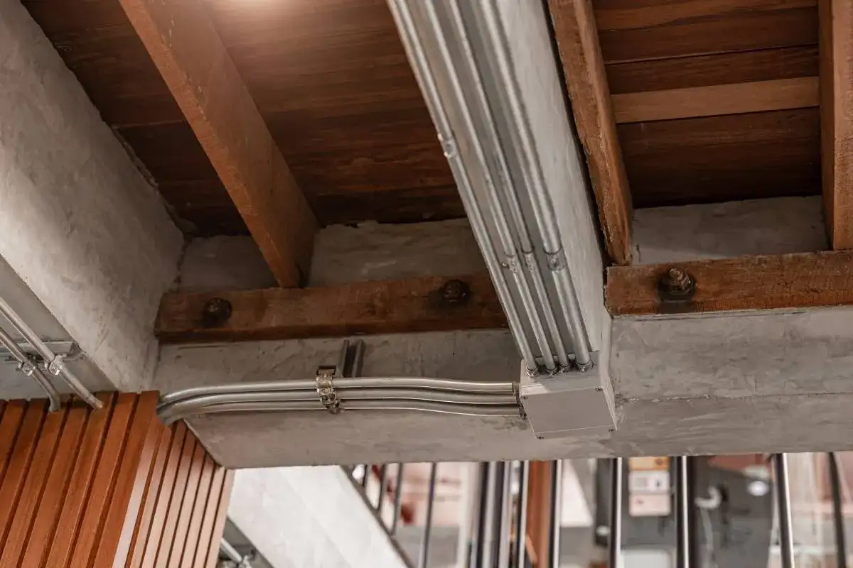 Metalowe rury elektryczne biegną wzdłuż betonowego podciągu, łącząc się w puszce. Drewniane belki stropowe i beton tworzą surową, industrialną estetykę.