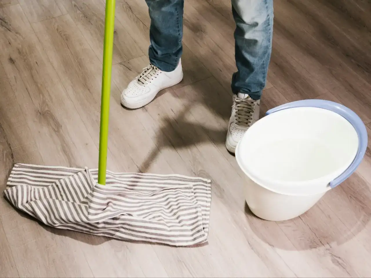 Osoba w jeansach i białych sneakersach przygotowuje się do mycia paneli. Obok mopa z pasiatą nakładką stoi wiadro z wodą.