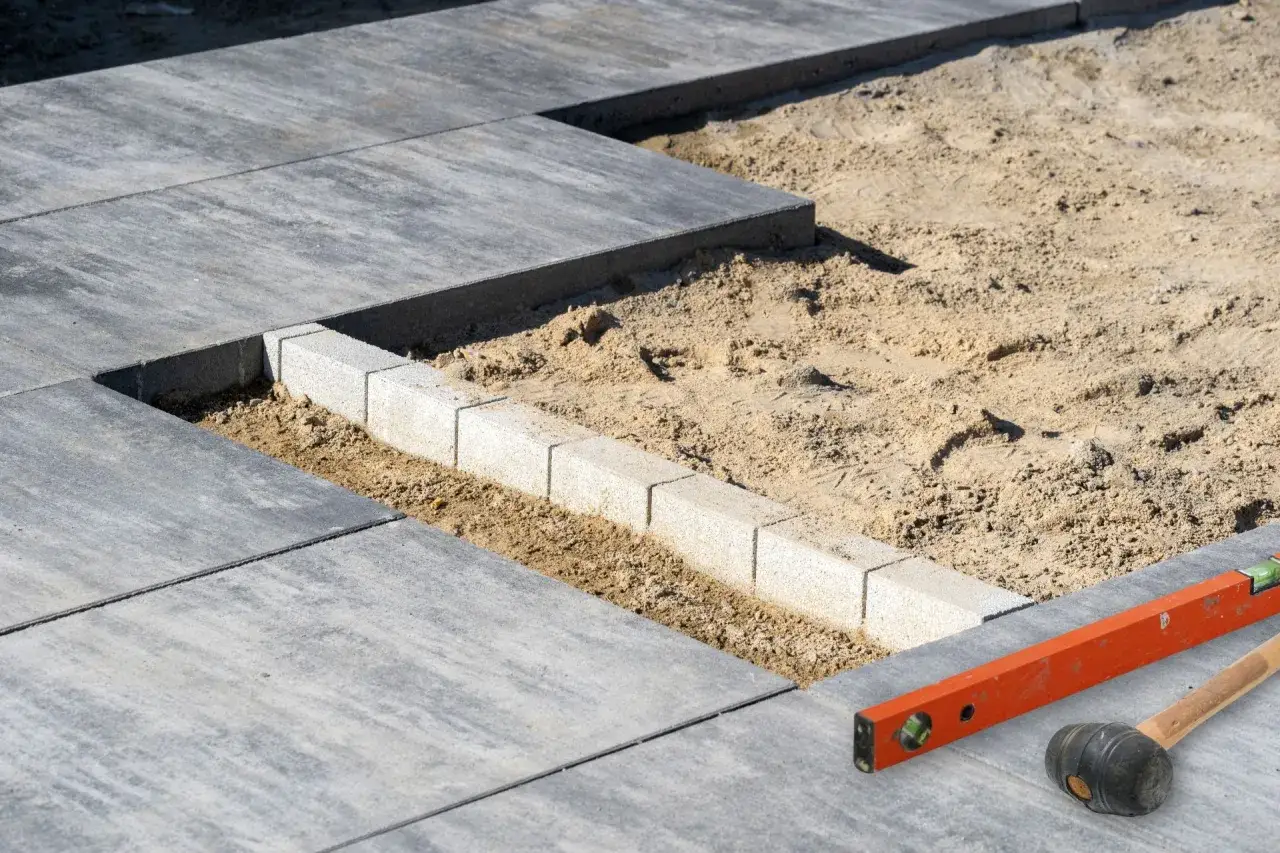 Układanie płyt na tarasie betonowym. Widać piasek, obrzeża i narzędzia: poziomicę i gumowy młotek.