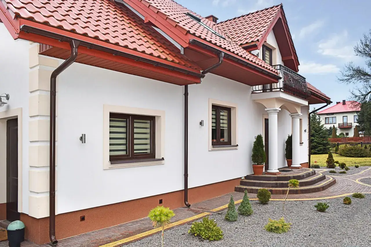 Elegancki dom z białym tynkiem, czerwonym dachem i ozdobnymi schodami. Podpowiadamy, jak zrobić tynk mozaikowy, by uzyskać podobny efekt.