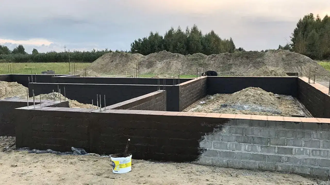 Fundament domu w budowie, ściany z bloczków betonowych pokryte ciemną izolacją. Piasek i zbrojenie czekają na dalsze prace.