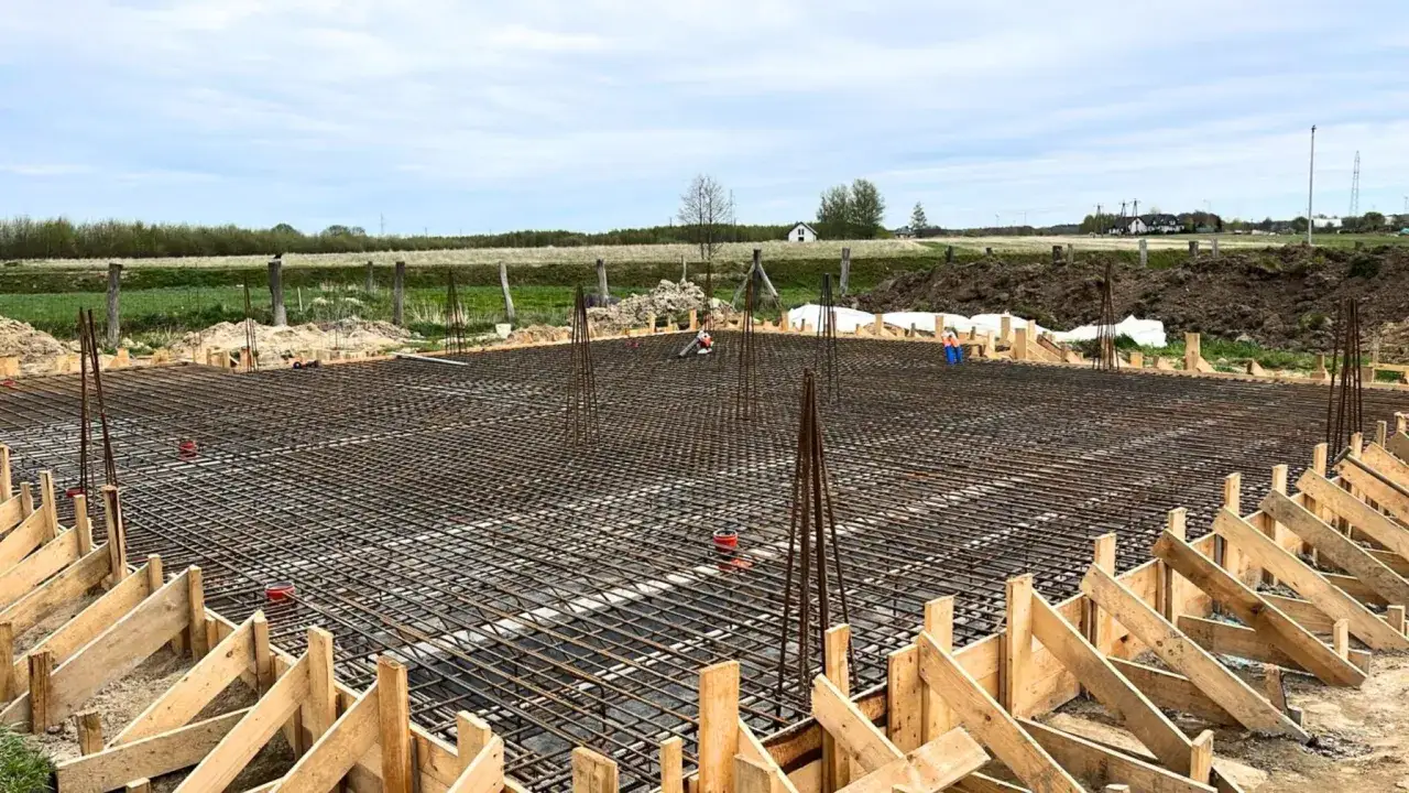 Fundament zbrojony i szalunki to początek budowy domu krok po kroku. W tle widać plac budowy i domy.