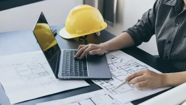 Architekt pracuje nad projektem, analizując plany i obliczając powierzchnię całkowitą budynku. Na biurku leży żółty kask.