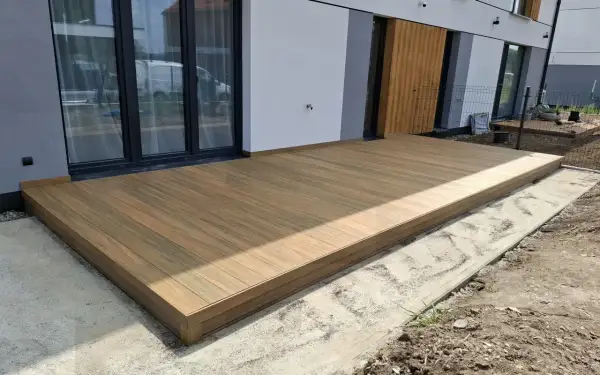 Nowy taras na słupach betonowych, z desek kompozytowych, przylega do nowoczesnego domu z dużymi przeszkleniami.