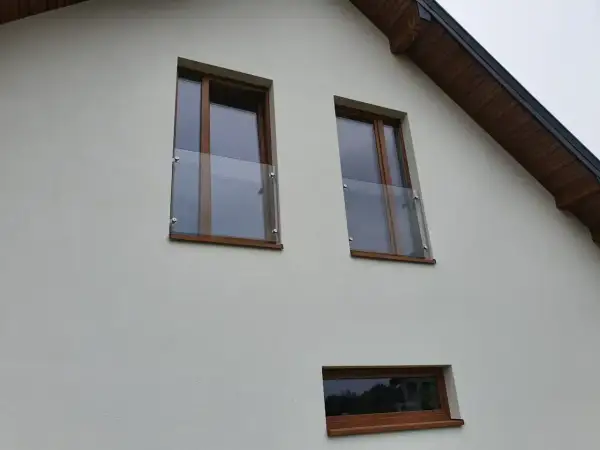 Nowoczesny portfenetr z drewnianymi ramami i szklaną balustradą przy oknach na białej elewacji.