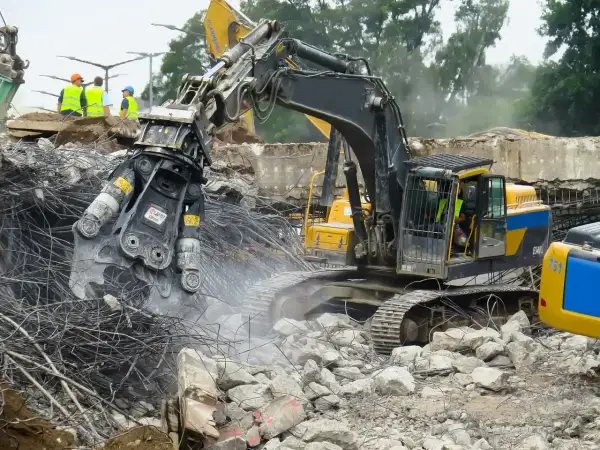 Maszyna budowlana wykonuje rozbiórkę budynku, krusząc beton i stal. W tle widać pracowników w kamizelkach odblaskowych.