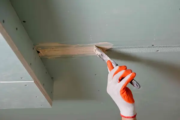 Ręka w rękawicy nakłada masę szpachlową na połączenie płyt gipsowo-kartonowych, przygotowując je do dalszych prac.